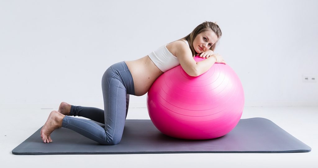 A pregnant woman leaning on a pink exercise ball.