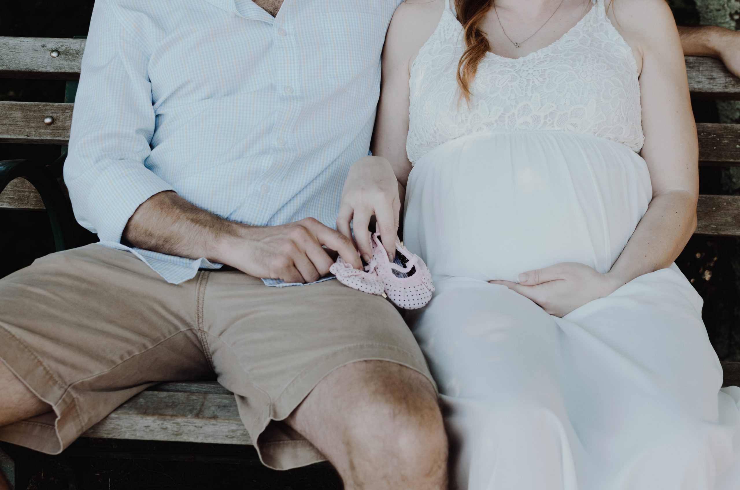 Torso of a pregnant couple sitting on the bench with children's shoes in their laps.