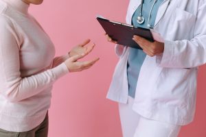 Ärztin unterhält sich stehend mit Patientin vor rosa Hintergrund.