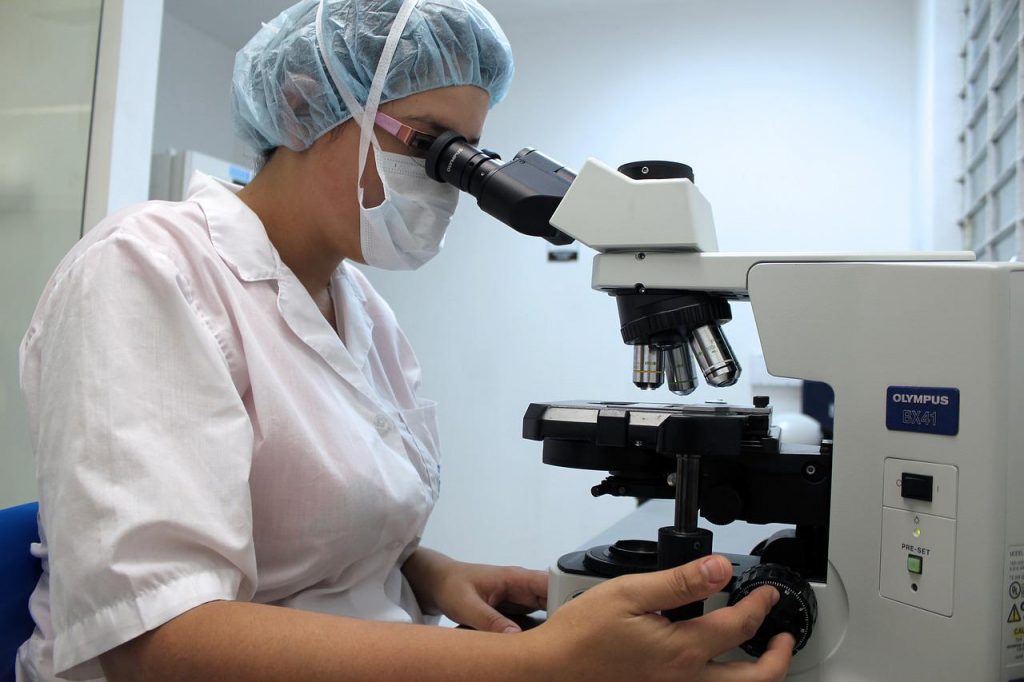 Woman in lab clothes uses a microscope while seated.