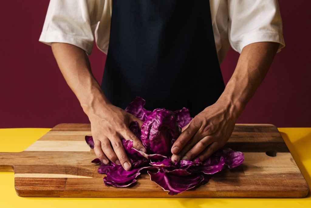 Torso of a man in a white T-shirt and blue apron in front of a crimson background and a yellow table on which a wooden board with pink cabbage leaves held in two hands lies.